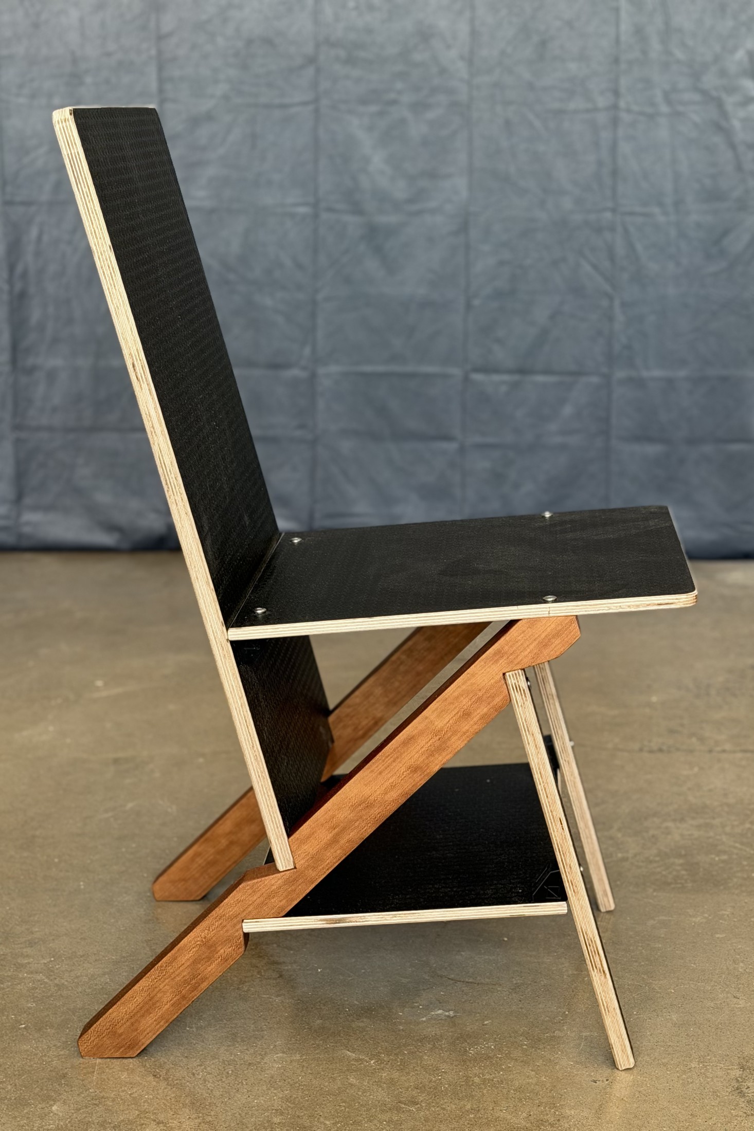 The chair in pure side elevation against a gray backdrop: black hexagonal-face plywood back, seat and tie panels seen edge-on with their pale ply edges showing, a cherry keel running as one long diagonal from the top of the back down to the floor, and a pale plywood front leg crossing it. The two triangles the chair is made of are visible as triangles.