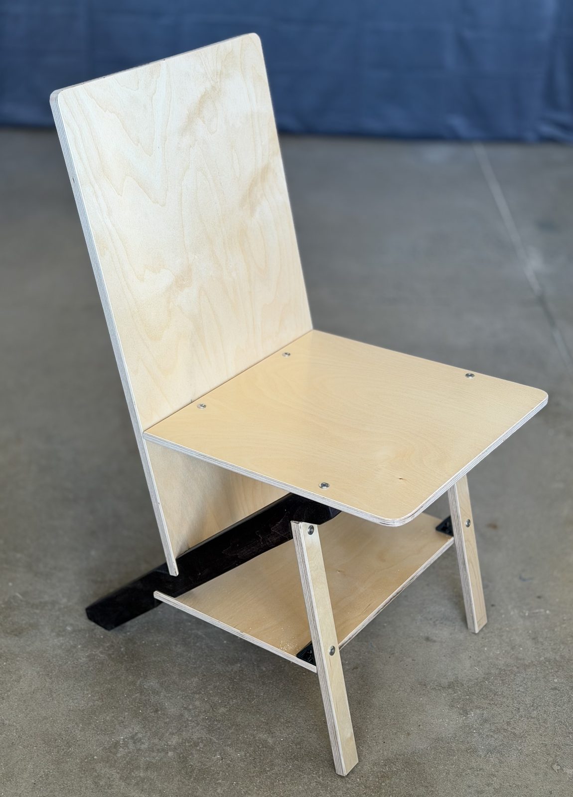 A single chair on a concrete floor against a blue backdrop, seen from the front and a little above: pale plywood back and seat panels with rounded corners and layered edges, two narrow plywood front legs with a plywood tie low between them, and a black keel running under the seat and back to the floor.