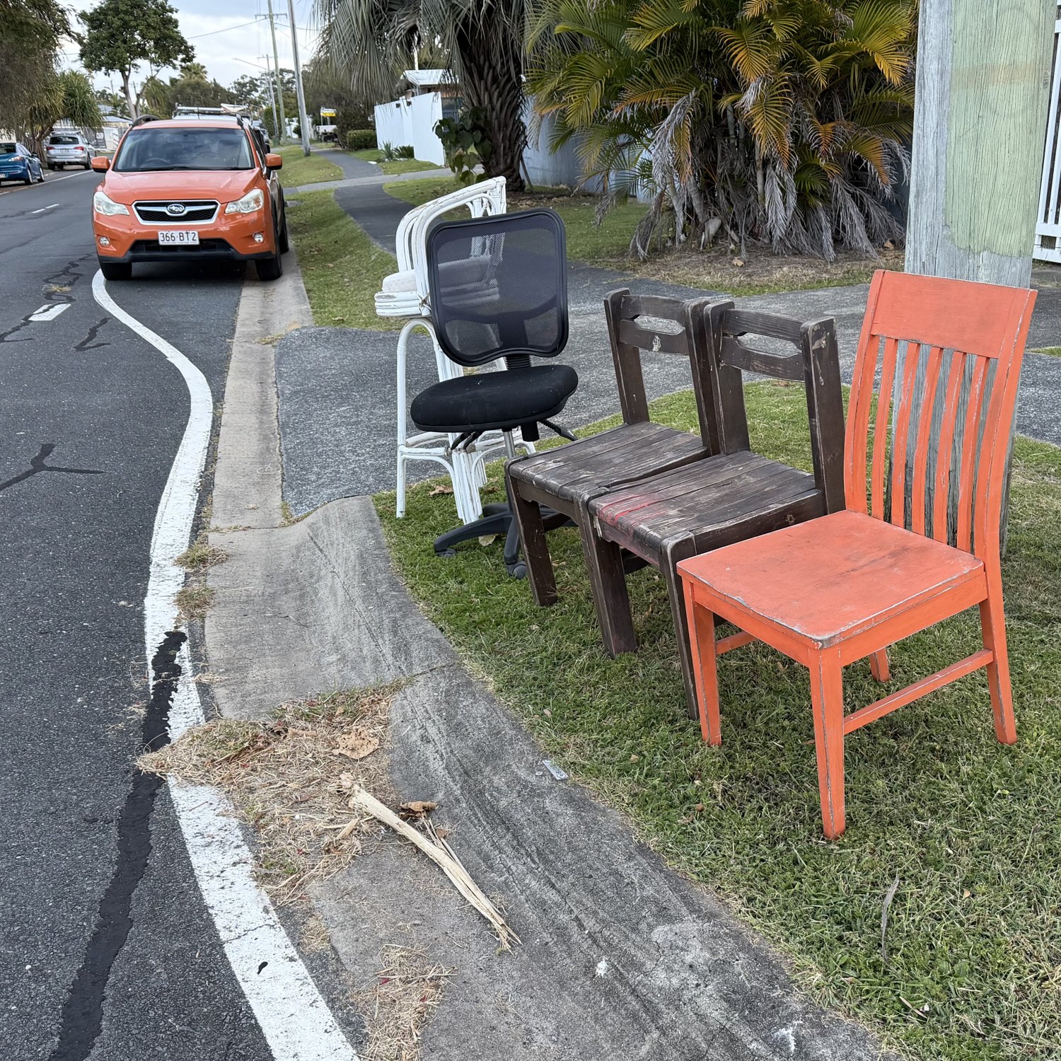 Four chairs set out on a suburban verge at dusk; an orange slat-back library chair at the end of the row.