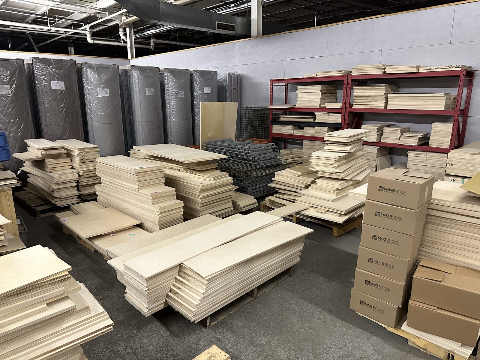 Stacks of pale birch plywood offcuts on pallets and red steel shelving in a warehouse, with MakerStock shipping boxes at right.