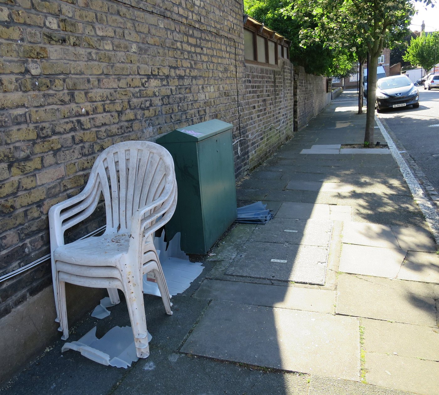 Three white plastic monobloc chairs, cracked and stacked, dumped against a brick wall on a London pavement