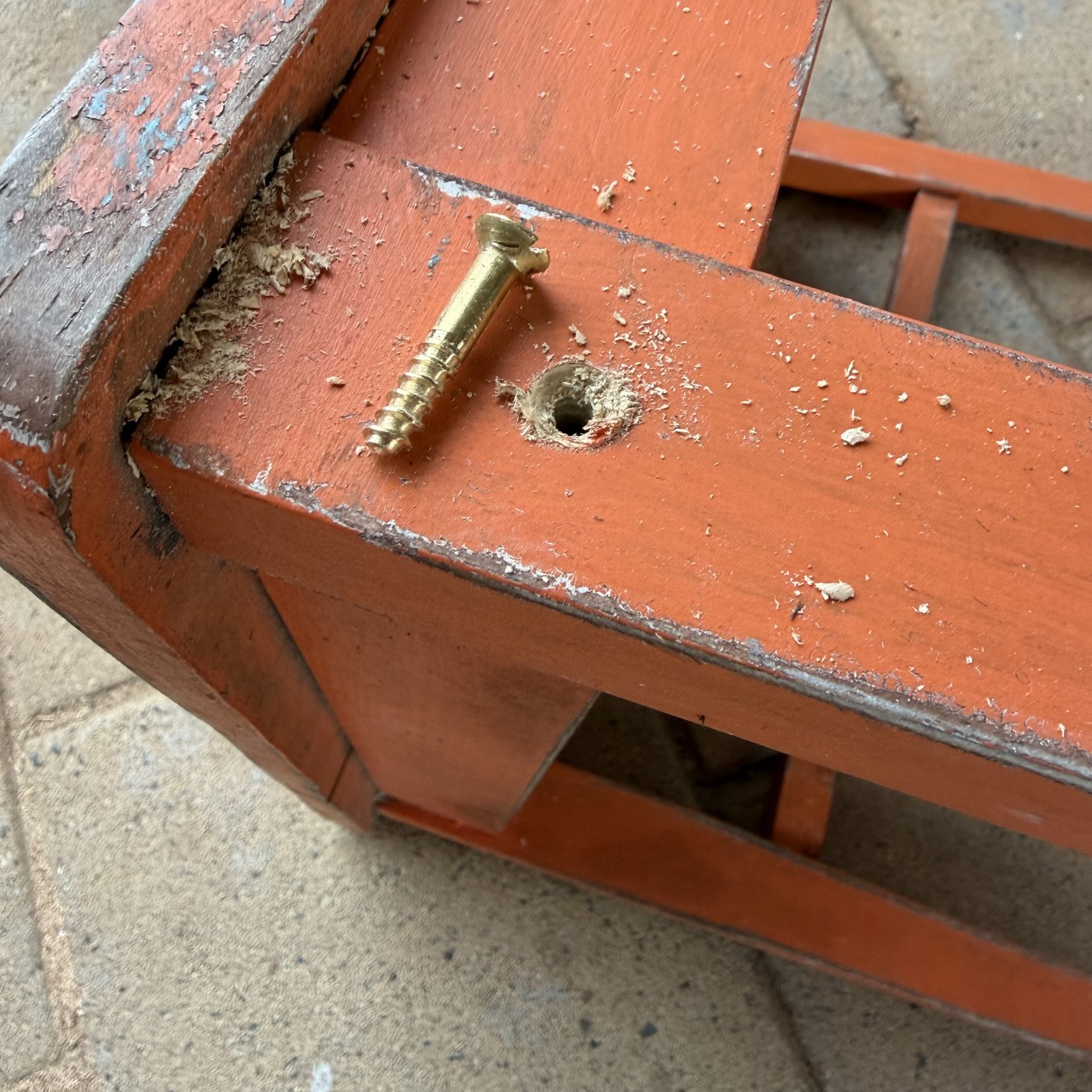 A broken brass wood screw lying on the orange-painted seat rail beside the hole it came out of. The countersunk head is torn open on one side and the lower end stops in a blunt, ragged fracture instead of a point. Fresh pale shavings are scattered across the rail, and where the paint has chipped along the edge a coat of pale blue shows under the orange.