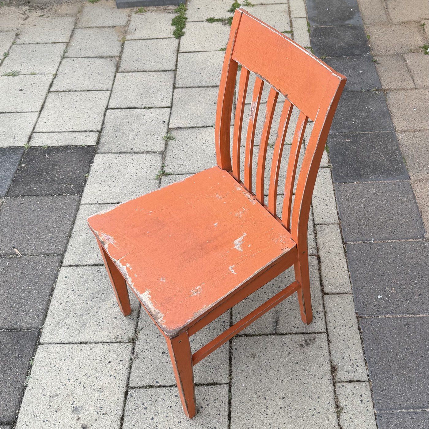 The orange chair cleaned up and standing on paving, photographed from above and to one side: a plain slat-back library chair with a curved crest rail, five vertical slats, a solid seat, square tapered legs and side stretchers. The orange paint is worn through to bare wood along the front edge of the seat and down the front legs.