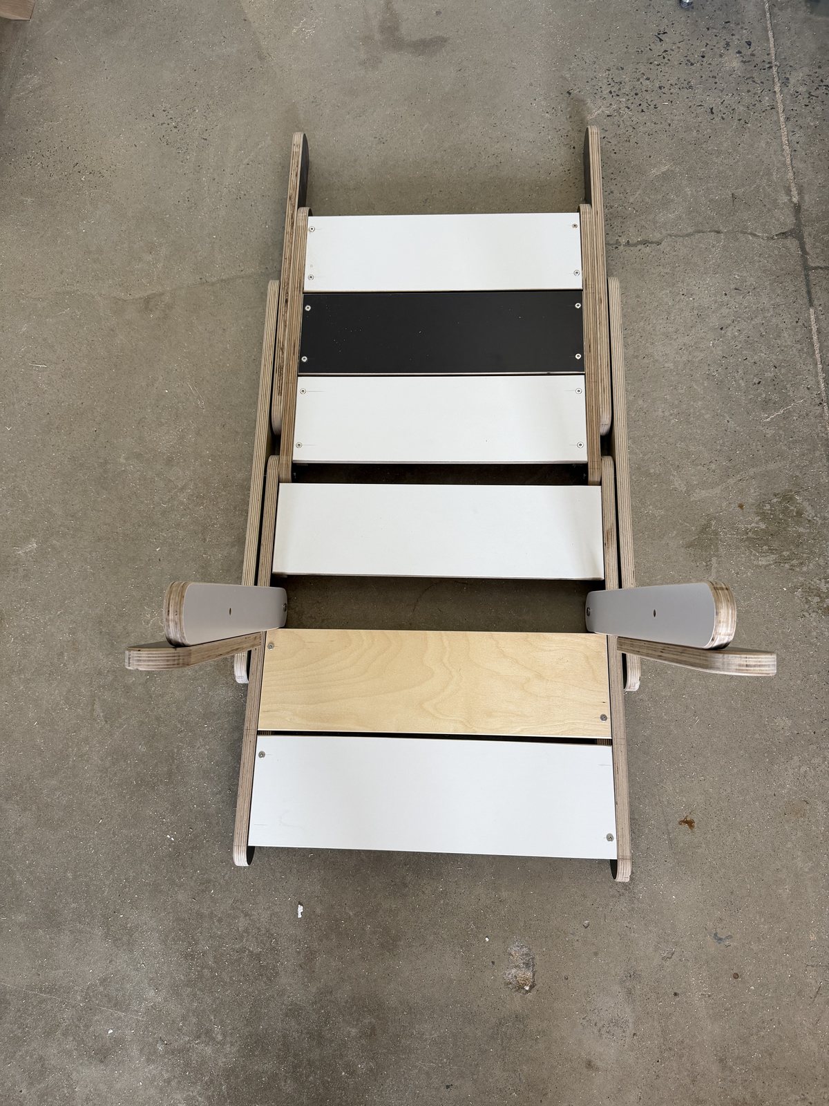 The third build, folded flat on the shop floor with the keel pin pulled: sticks lying parallel, panels stacked, the two arms standing out sideways because the back panel is in their way.