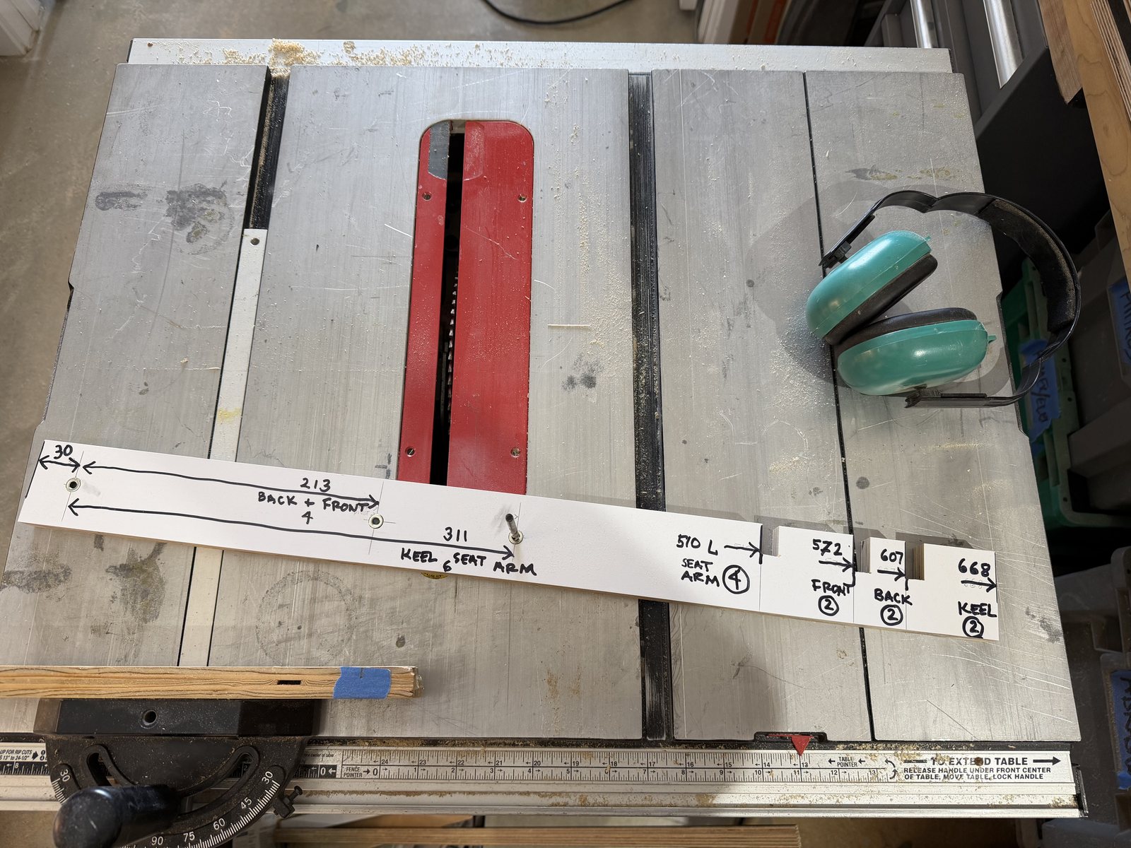 The story pole lying on the table saw: a plywood strip with three drill positions marked on one half, 30, 213 and 311, and four notched length stops on the other, 510, 572, 607 and 668, each labeled in pencil with the part it cuts.