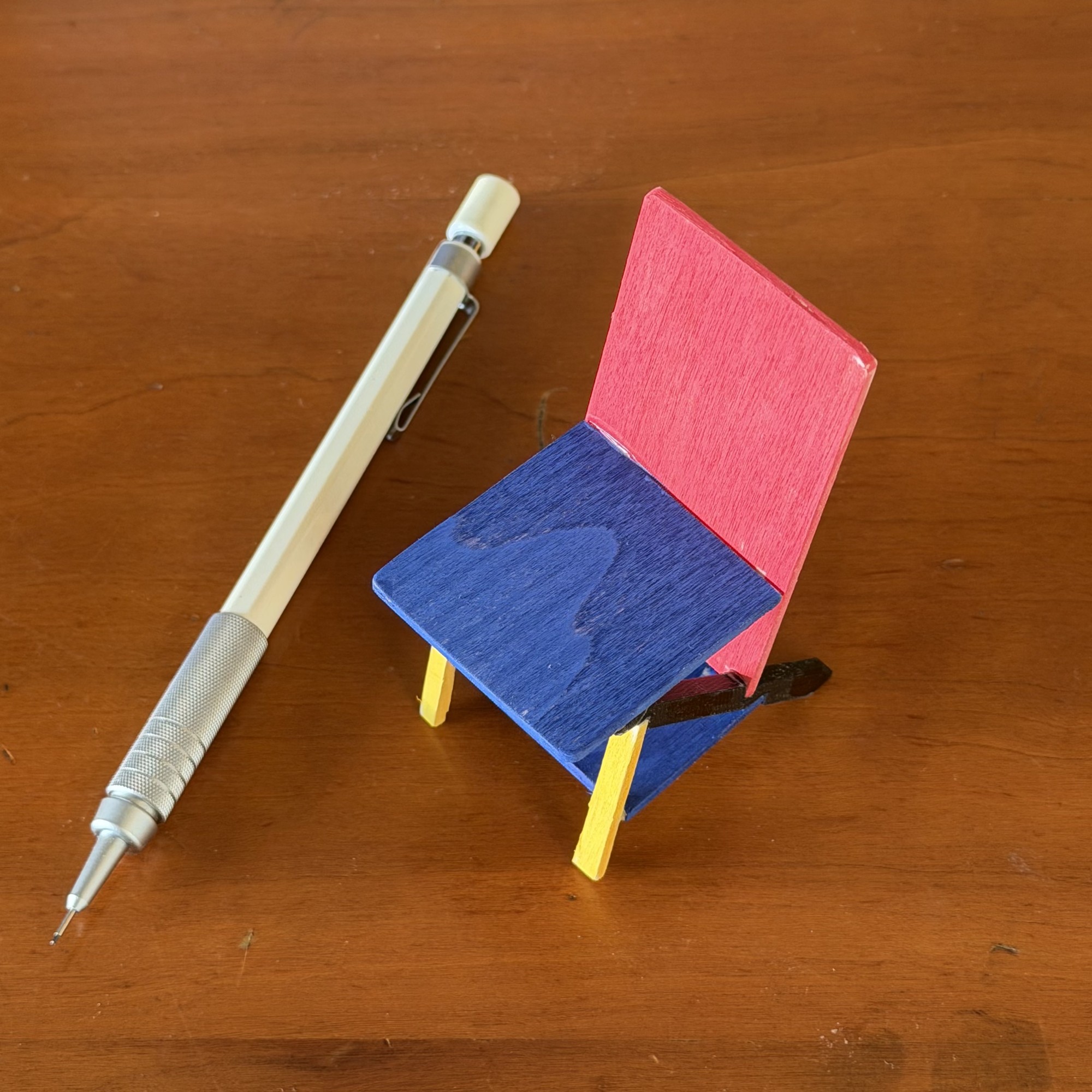 A chair model at one tenth scale standing on a wooden table beside a mechanical pencil for scale, reaching roughly half the pencil's length. The back panel is red, the seat and the tie panel below it are blue, the keel running under the seat is black, and the two front legs are yellow.