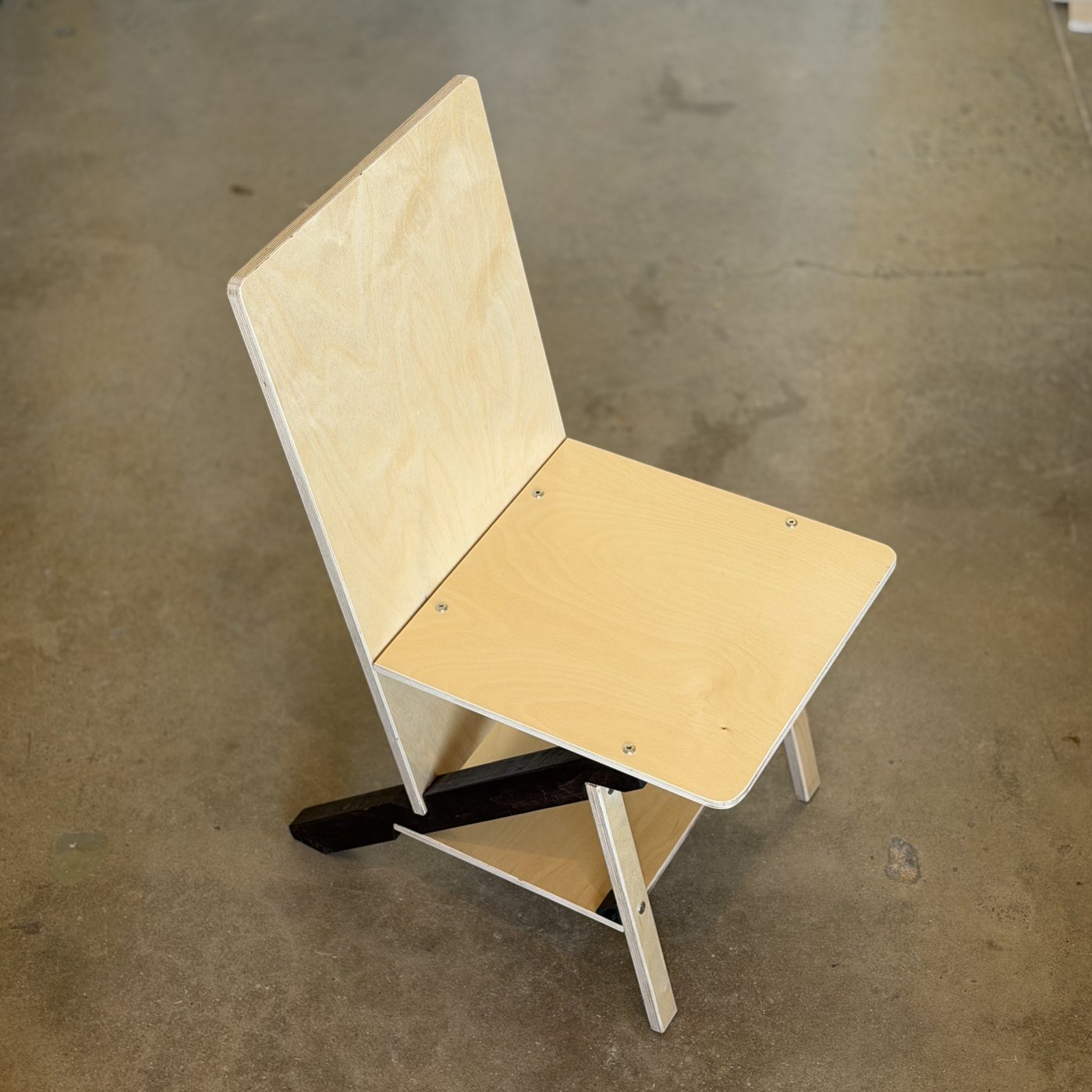 Cherry 1 from the front quarter: birch plywood back, seat, front and tie on a dark cherry keel, the seat carried on four recessed furniture bolts, every edge showing its pale ply lines.
