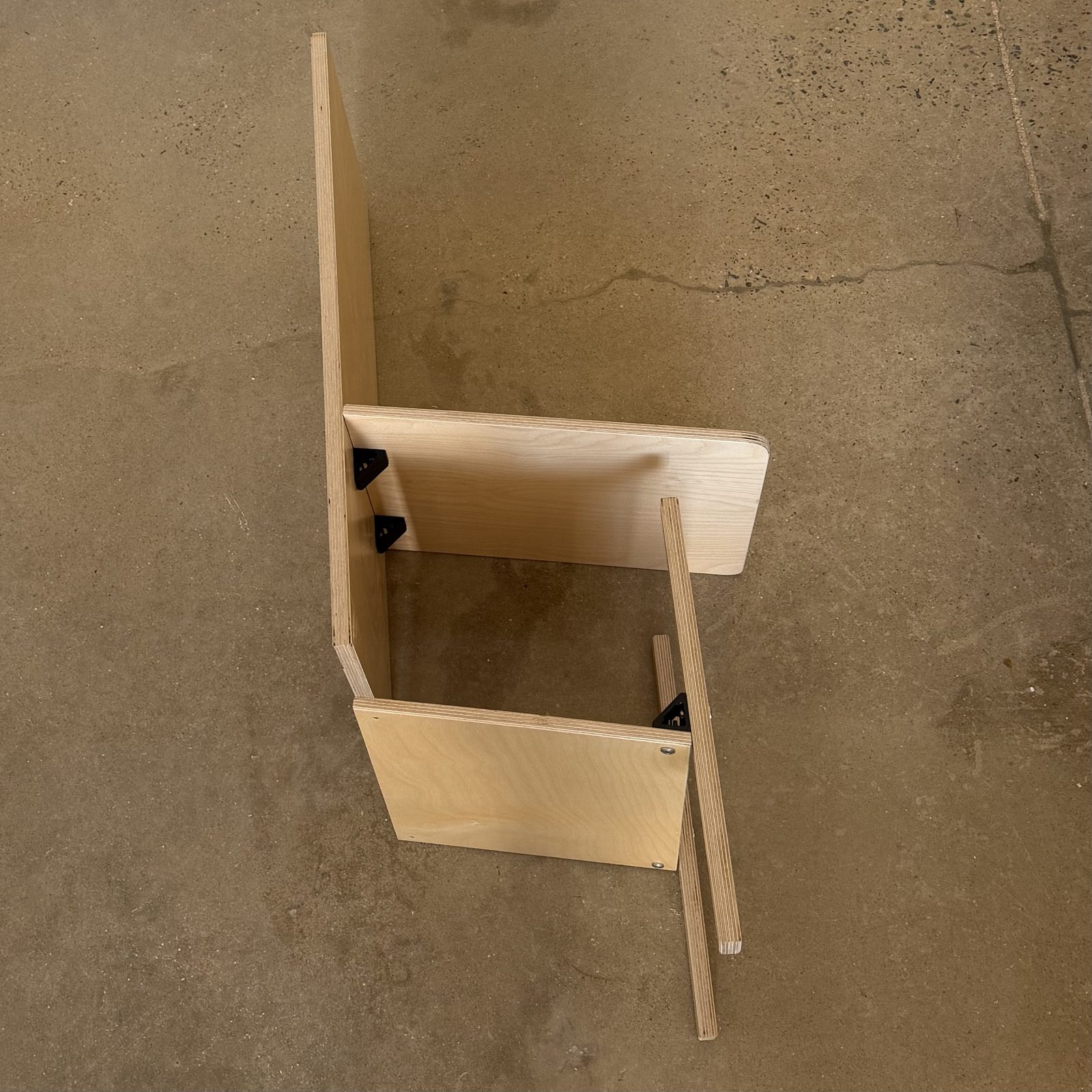 A Keel Panel part-assembled on the shop floor: back, seat, front and tie panels in pale birch plywood standing at their finished angles, held only by small black brackets, with the keel not yet fitted and the tie stick lying loose alongside.