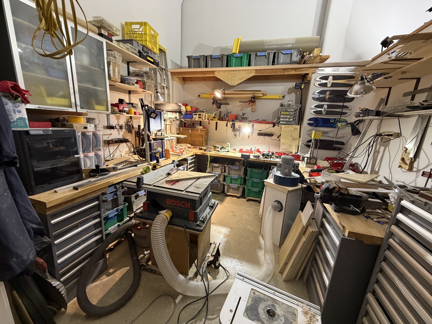 The shop at the end of the design phase: a portable table saw and a dust collector out in the middle of a small floor, benches and tool chests around three walls, offcuts stacked on end, skateboard decks racked overhead.
