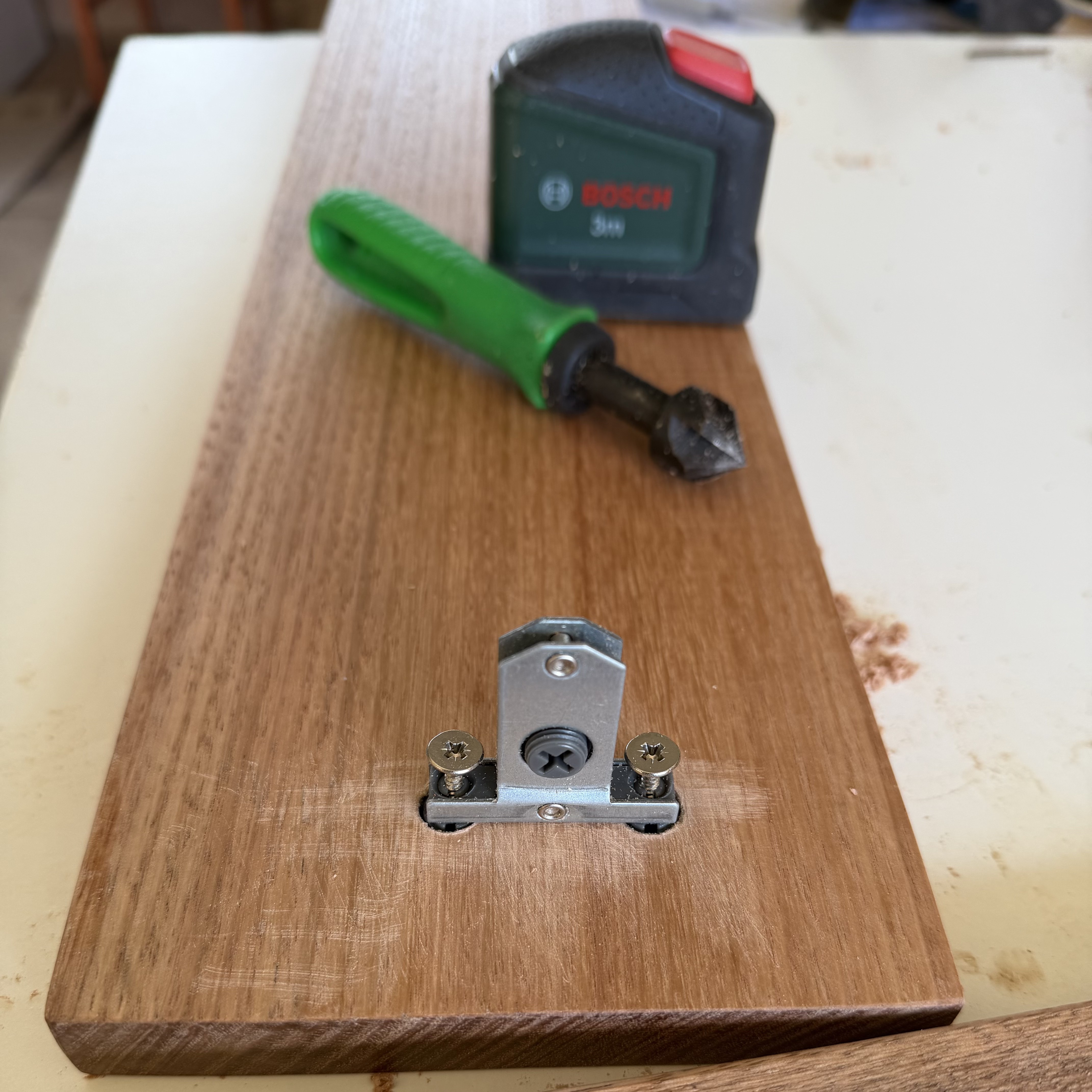 A zinc-plated steel bracket shaped like a low house, set flush into the end of an oak board. A central cross-head adjuster screw sits in a black plastic collar, and a cross-head screw on each side is driven into a recessed pocket in the wood; a black plastic channel runs along the bracket's base to clip onto the drawer's side rail. A green-handled countersink and a Bosch tape measure lie on the bench behind.