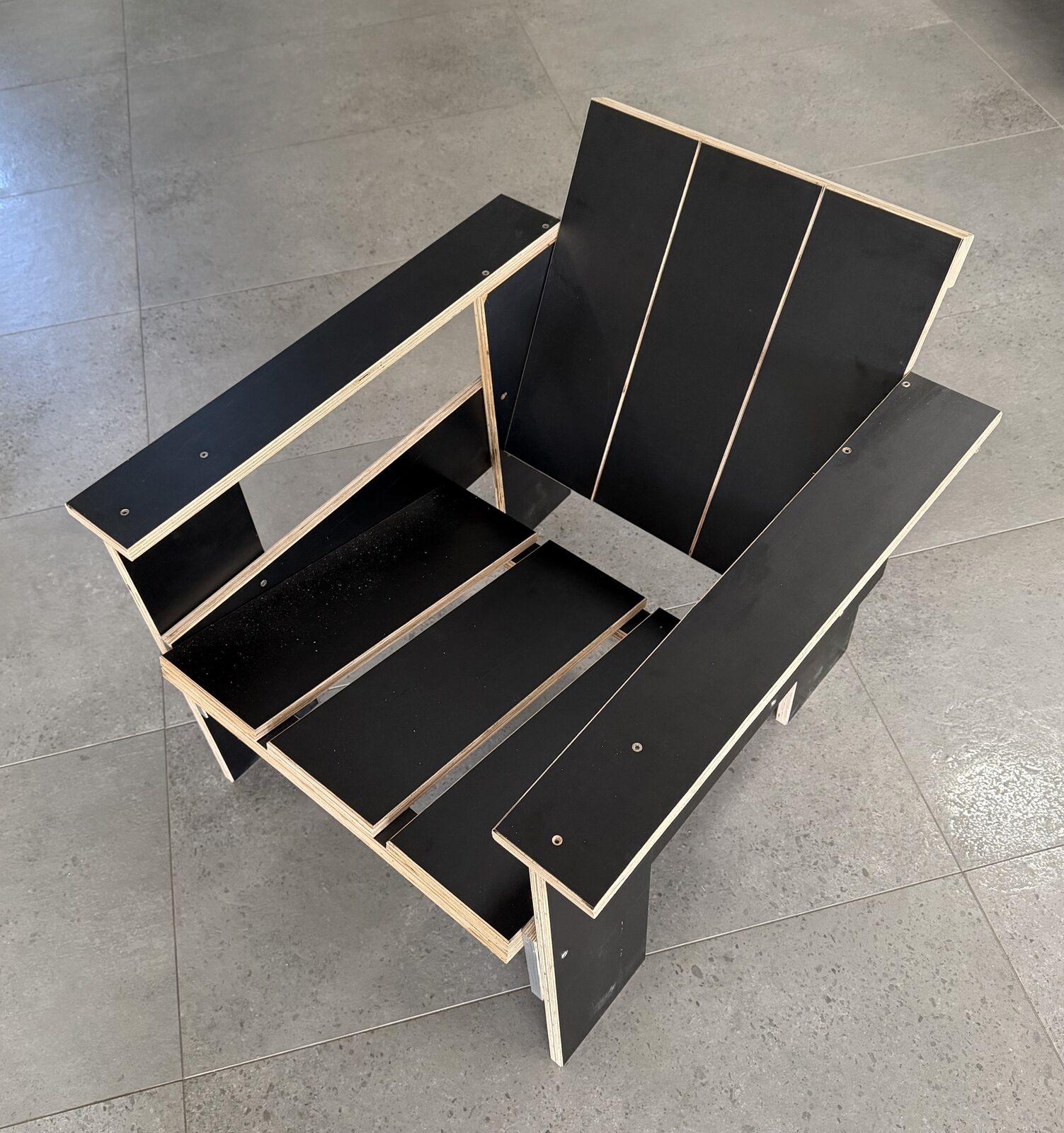 The crate lounge chair rebuilt in black film-faced plywood: slatted back and seat, wide flat arms, pale edge plies outlining every board.